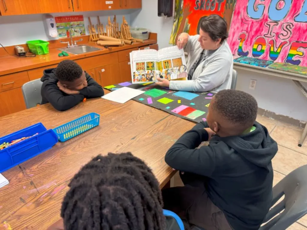 A woman reads a colorful book aloud to three attentive children at a table, surrounded by art supplies in a bright classroom