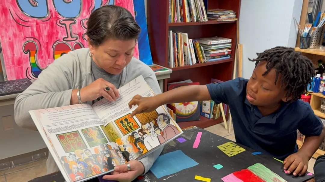 A child points at an open storybook while an adult reads, surrounded by colorful materials in a cozy, creative environment.